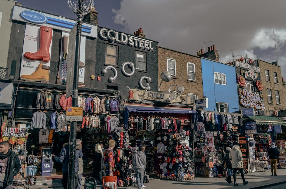 A street view of Camden Market, featuring a row of vibrant, multi-colored market stalls with clothing, hats, and accessories displayed outside. The storefronts are housed in brick buildings, some with