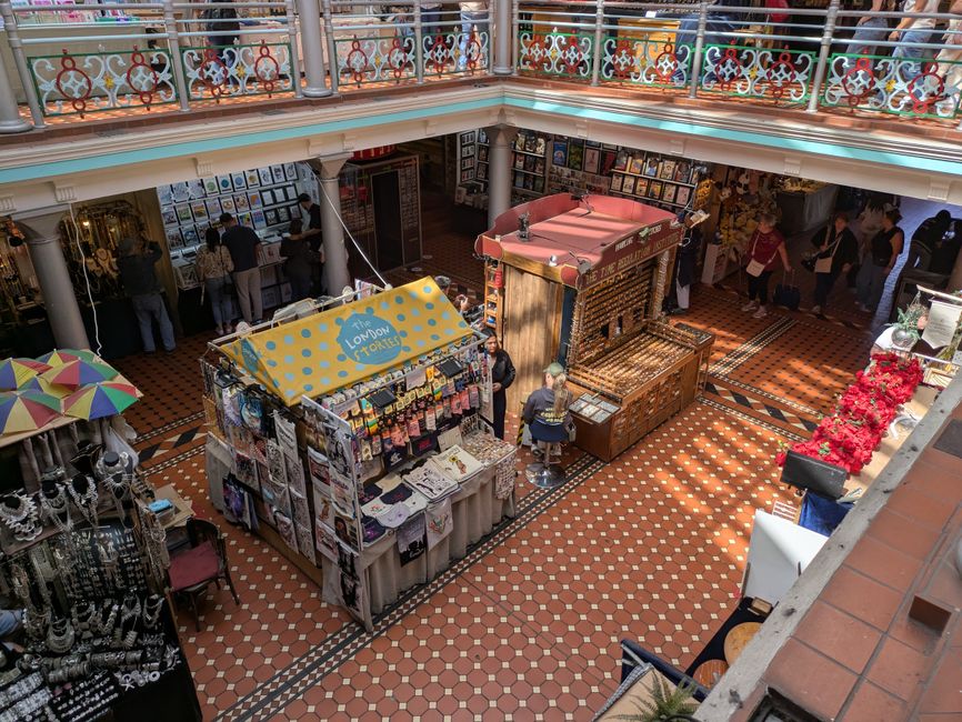 A street view of Camden Market, featuring a row of vibrant, multi-colored market stalls with clothing, hats, and accessories displayed outside. The storefronts are housed in brick buildings, some with