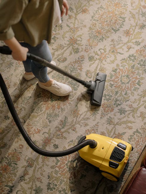 A professional cleaning technician dressed in a white protective suit, face mask, gloves, and goggles is performing a deep cleaning or sanitisation process on a beige carpet within a modern, well-lit