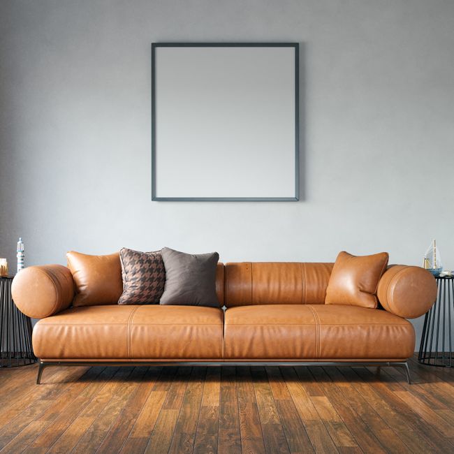 Close-up of a light grey upholstered armchair with a rounded armrest and wooden legs, situated on a wooden floor next to sheer white curtains that diffuse natural daylight into a modern domestic setti